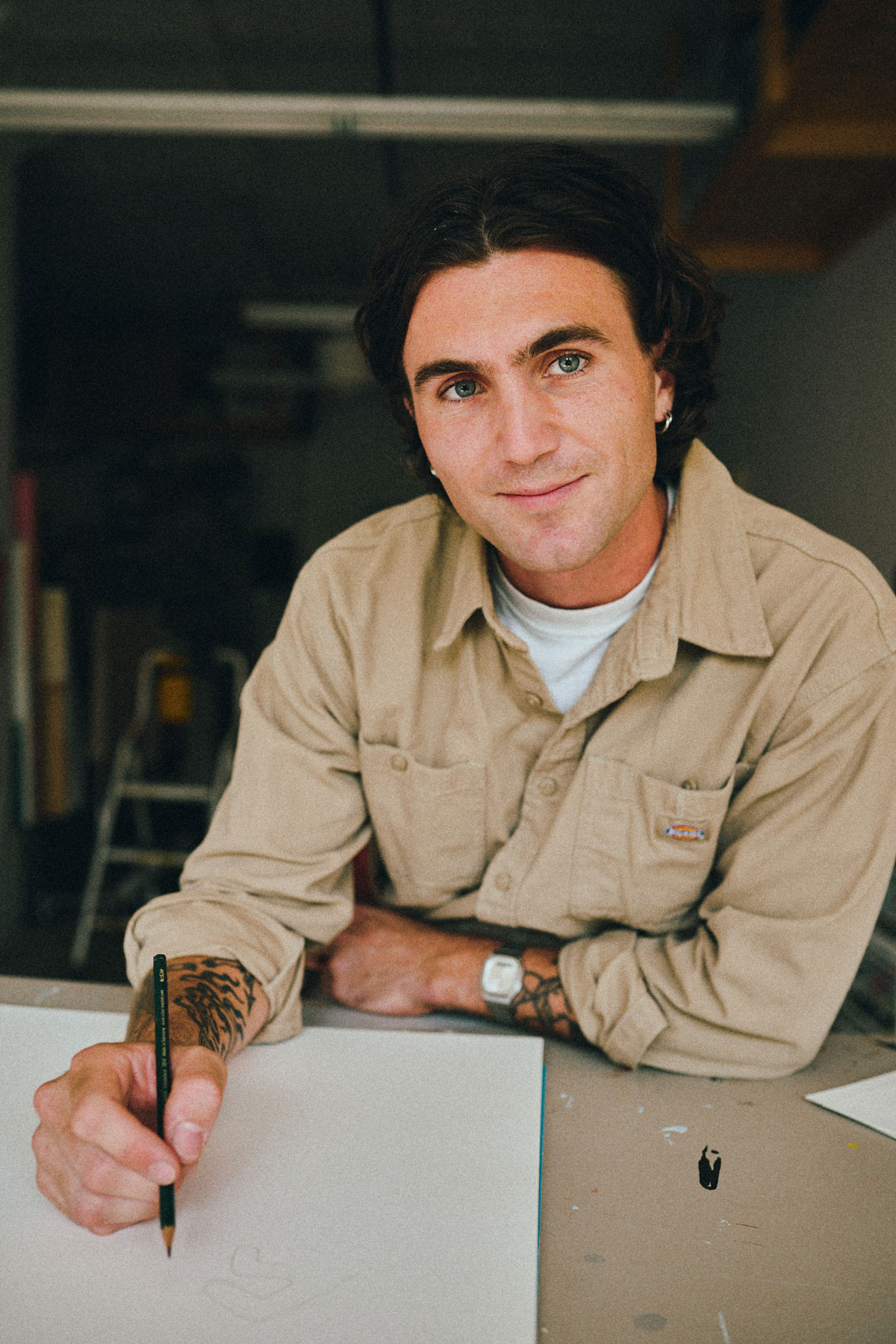Portrait of Oliver Gadoury at a studio table, smiling at the camera with a pencil on a sheet of paper.